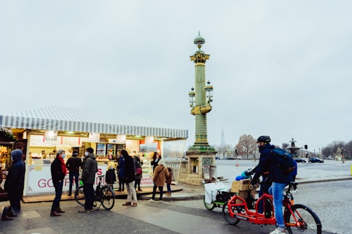 A group of people is gathered in front of a street food stall, possibly selling sandwiches and other snacks. The stall has a striped awning, and people appear to be queuing. A cyclist on a red bicycle is passing by, carrying goods in a cargo box on the back. A decorative street lamp stands nearby in an urban setting.