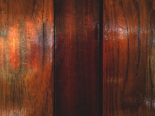 A close-up view of polished wooden panels with visible grain patterns and rich, warm colors reflecting light. The wood exhibits a glossy texture with areas of darker and lighter shades.