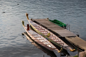 A long, decorative dragon boat is moored beside a wooden dock on calm water. The boat features intricate patterns and dragon-shaped ornaments at the front and rear. Next to the dragon boat is a small green rowboat. The water gently ripples with a few ducks swimming nearby, creating a peaceful scene.
