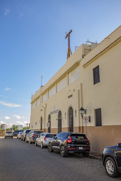 A street scene with several cars parked along a paved road next to a large beige building with ornate black iron doors and a logo reading 'Colegio Santa Clara' above one entrance. The building has a flat roof with visible utility structures, and the sky is bright blue with a few clouds.
