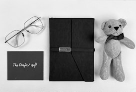 A neatly arranged collection of items displayed on a white textured surface includes a pair of eyeglasses with thin frames, a hardcover black notebook secured with a strap, a greeting card that reads 'The Perfect Gift' in white script, and a knitted teddy bear with a black bow around its neck.