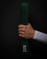 A hand holds a green martial arts belt against a dark background. The belt displays a patch with the word 'BLITZ' in white letters. The hand is clenched around the folded belt, showing visible veins and a light skin tone. The person is wearing a white long-sleeved garment.