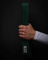 A hand holds a green martial arts belt against a dark background. The belt displays a patch with the word 'BLITZ' in white letters. The hand is clenched around the folded belt, showing visible veins and a light skin tone. The person is wearing a white long-sleeved garment.