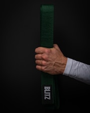 A hand holds a green martial arts belt against a dark background. The belt displays a patch with the word 'BLITZ' in white letters. The hand is clenched around the folded belt, showing visible veins and a light skin tone. The person is wearing a white long-sleeved garment.