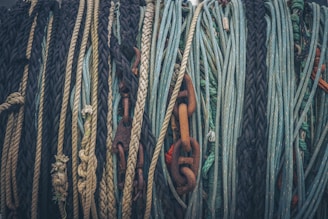 Multiple ropes and a rusty metal chain are tightly wound together. The ropes vary in color and texture, featuring shades of blue, green, brown, and black. The different textures create a complex and visually captivating pattern.