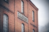 A red brick building with multiple tall, rectangular windows is shown. A sign that reads 'G.A. Jensen' is prominently displayed on the side of the building. The architecture of the structure appears industrial or historical, suggesting an older factory or warehouse. The sky is overcast, adding a dim gray backdrop to the scene.