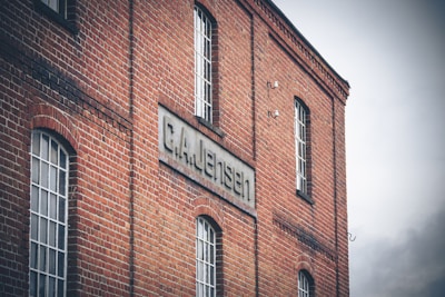 A red brick building with multiple tall, rectangular windows is shown. A sign that reads 'G.A. Jensen' is prominently displayed on the side of the building. The architecture of the structure appears industrial or historical, suggesting an older factory or warehouse. The sky is overcast, adding a dim gray backdrop to the scene.