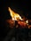Bright flames rise from a pile of burning wood, casting an orange and yellow glow against a dark background. The fire illuminates the surrounding area, highlighting the texture of the sticks and creating a warm, dynamic visual.