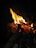 Bright flames rise from a pile of burning wood, casting an orange and yellow glow against a dark background. The fire illuminates the surrounding area, highlighting the texture of the sticks and creating a warm, dynamic visual.