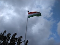 A flag is fluttering on a tall flagpole against a backdrop of a cloudy sky. The flag features three horizontal stripes of saffron, white, and green with a blue wheel in the center of the white stripe. Dark green trees are visible at the bottom, partially silhouetted against the sky.