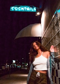 A woman stands confidently on a dimly lit street at night, leaning against a tiled wall under a glowing neon sign that reads 'Paradise Cocktails'. She is wearing a white tank top, camo pants, and a denim jacket, with a relaxed pose.