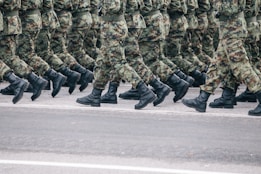 A group of soldiers march in unison wearing camouflage uniforms and black boots. The soldiers are aligned in rows, showing discipline and uniformity as they move along a paved road. The focus is on their lower bodies, highlighting the precision of their steps.