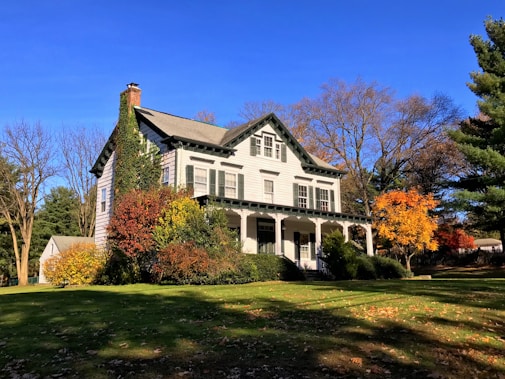 A charming two-story house with white siding and dark green shutters sits amidst a lush garden. The house is surrounded by trees with autumn foliage in vibrant shades of orange, red, and yellow. Ivy climbs up part of the house, adding to the picturesque setting. A well-maintained lawn stretches in front of the house under a clear blue sky, and there is a smaller building visible in the background on the left side.