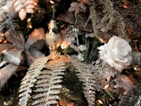 A festive Christmas decoration featuring a variety of elements including a large silver bow, a white rose, glittering fern leaves, and other ornamental foliage. There are warm white string lights intertwined with the decorations, creating a cozy and celebratory atmosphere.