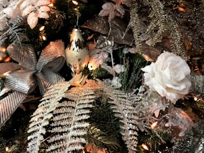A festive Christmas decoration featuring a variety of elements including a large silver bow, a white rose, glittering fern leaves, and other ornamental foliage. There are warm white string lights intertwined with the decorations, creating a cozy and celebratory atmosphere.