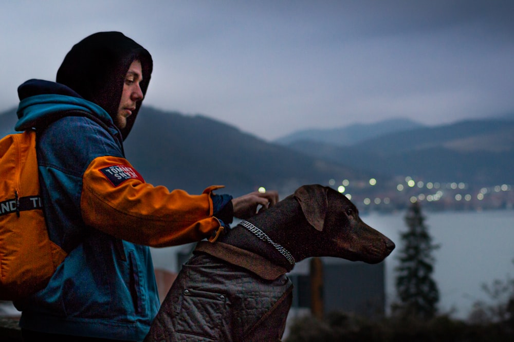 man in blue jacket and black hat holding black labrador retriever during daytime