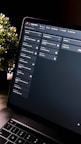 A close-up view of a laptop screen displaying a project management tool with columns labeled 'Open', 'In Progress', and 'Review'. Tasks are listed under each column. The environment appears dimly lit, with a small, leafy plant visible in the background.