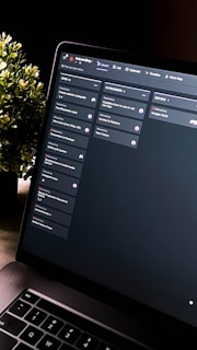 A close-up view of a laptop screen displaying a project management tool with columns labeled 'Open', 'In Progress', and 'Review'. Tasks are listed under each column. The environment appears dimly lit, with a small, leafy plant visible in the background.