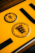 A close-up of a yellow vehicle registration plate displaying black symbols and text. The upper section features a circular inspection sticker with numerical markings from 1 to 12 and a highlighted number 10 in the center. Below, a circular emblem contains the text 'Baden-Württemberg' and a stylized coat of arms with lions.