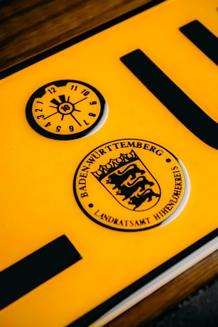 A close-up of a yellow vehicle registration plate displaying black symbols and text. The upper section features a circular inspection sticker with numerical markings from 1 to 12 and a highlighted number 10 in the center. Below, a circular emblem contains the text 'Baden-Württemberg' and a stylized coat of arms with lions.