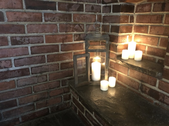 A rustic brick corner featuring a collection of white candles on dark stone shelves. A large metallic lantern with an open door contains a lit candle, casting a warm glow. The bricks have a worn, textured appearance, contributing to a cozy, intimate setting.