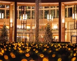 A decorated outdoor space with a large structure draped in festive lights, surrounded by glowing bokeh effects. Nearby, there are trees and a building with illuminated interiors and large windows. Hanging pendant lights add to the warm and inviting ambiance.