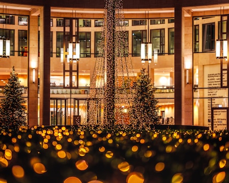 A decorated outdoor space with a large structure draped in festive lights, surrounded by glowing bokeh effects. Nearby, there are trees and a building with illuminated interiors and large windows. Hanging pendant lights add to the warm and inviting ambiance.