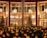 A decorated outdoor space with a large structure draped in festive lights, surrounded by glowing bokeh effects. Nearby, there are trees and a building with illuminated interiors and large windows. Hanging pendant lights add to the warm and inviting ambiance.