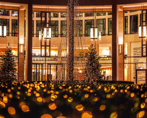 A decorated outdoor space with a large structure draped in festive lights, surrounded by glowing bokeh effects. Nearby, there are trees and a building with illuminated interiors and large windows. Hanging pendant lights add to the warm and inviting ambiance.