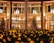 A decorated outdoor space with a large structure draped in festive lights, surrounded by glowing bokeh effects. Nearby, there are trees and a building with illuminated interiors and large windows. Hanging pendant lights add to the warm and inviting ambiance.