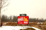A bright red sign is displayed in an open, snowy field with bare trees in the background. The sign warns about fines for failure to comply with COVID-19 regulations, with visible text including a URL for more information. The scene appears overcast, with muted natural colors.