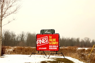 A bright red sign is displayed in an open, snowy field with bare trees in the background. The sign warns about fines for failure to comply with COVID-19 regulations, with visible text including a URL for more information. The scene appears overcast, with muted natural colors.