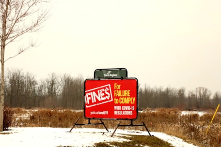 A bright red sign is displayed in an open, snowy field with bare trees in the background. The sign warns about fines for failure to comply with COVID-19 regulations, with visible text including a URL for more information. The scene appears overcast, with muted natural colors.