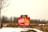 A bright red sign is displayed in an open, snowy field with bare trees in the background. The sign warns about fines for failure to comply with COVID-19 regulations, with visible text including a URL for more information. The scene appears overcast, with muted natural colors.