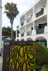 A decorative sign with the text 'GRACIAS & ADIOS' in colorful, ornate lettering set against a backdrop of a white building with balconies. A palm tree stands beside the building, and there are green plants in the foreground. The sky appears partly cloudy, adding to the relaxed, tropical atmosphere.
