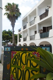 A decorative sign with the text 'GRACIAS & ADIOS' in colorful, ornate lettering set against a backdrop of a white building with balconies. A palm tree stands beside the building, and there are green plants in the foreground. The sky appears partly cloudy, adding to the relaxed, tropical atmosphere.