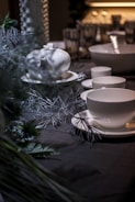 A dimly lit table setting features a white teapot and teacups on saucers. The tablecloth is dark, and there are decorative elements like branches and foliage. Glasses and bowls are visible in the background, hinting at a sophisticated setup.