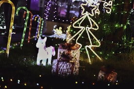 A festive outdoor setting features illuminated decorations, including candy cane lights lining a path, a glowing unicorn figure, a wire-frame Christmas tree with a star on top, and several wrapped gift boxes. The scene is adorned with colorful string lights, creating a bright and cheerful atmosphere.