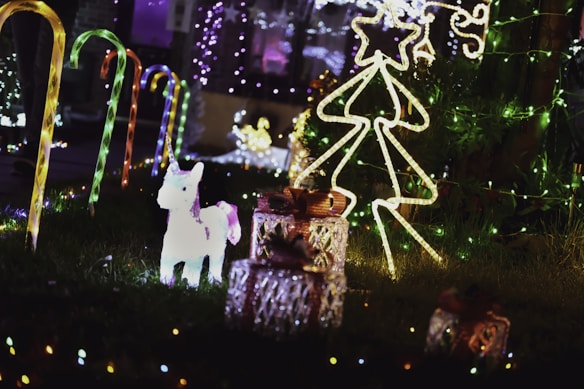 A festive outdoor setting features illuminated decorations, including candy cane lights lining a path, a glowing unicorn figure, a wire-frame Christmas tree with a star on top, and several wrapped gift boxes. The scene is adorned with colorful string lights, creating a bright and cheerful atmosphere.