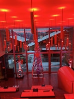 A modern interior space features a vibrant red lighting scheme with long cylindrical lights hanging from the ceiling. There is a large window overlooking a snowy landscape with buildings. Inside, a metal sculpture stands next to other art pieces and sleek contemporary furniture.