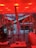 A modern interior space features a vibrant red lighting scheme with long cylindrical lights hanging from the ceiling. There is a large window overlooking a snowy landscape with buildings. Inside, a metal sculpture stands next to other art pieces and sleek contemporary furniture.