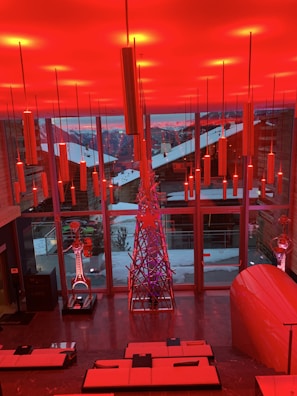 A modern interior space features a vibrant red lighting scheme with long cylindrical lights hanging from the ceiling. There is a large window overlooking a snowy landscape with buildings. Inside, a metal sculpture stands next to other art pieces and sleek contemporary furniture.