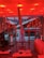 A modern interior space features a vibrant red lighting scheme with long cylindrical lights hanging from the ceiling. There is a large window overlooking a snowy landscape with buildings. Inside, a metal sculpture stands next to other art pieces and sleek contemporary furniture.