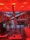 A modern interior space features a vibrant red lighting scheme with long cylindrical lights hanging from the ceiling. There is a large window overlooking a snowy landscape with buildings. Inside, a metal sculpture stands next to other art pieces and sleek contemporary furniture.