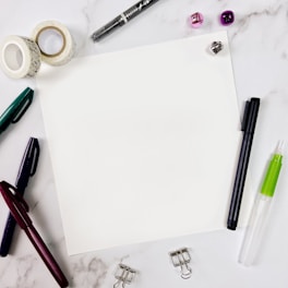 A blank sheet of paper sits in the center surrounded by various stationery items on a marble surface. Items include pens in different colors, decorative tape rolls, a black marker, a green and white glue pen, a transparent adhesive dispenser, clip holders, and small shiny jingle bells in purple and silver.