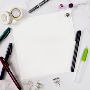 A blank sheet of paper sits in the center surrounded by various stationery items on a marble surface. Items include pens in different colors, decorative tape rolls, a black marker, a green and white glue pen, a transparent adhesive dispenser, clip holders, and small shiny jingle bells in purple and silver.