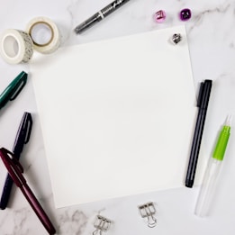 A blank sheet of paper sits in the center surrounded by various stationery items on a marble surface. Items include pens in different colors, decorative tape rolls, a black marker, a green and white glue pen, a transparent adhesive dispenser, clip holders, and small shiny jingle bells in purple and silver.