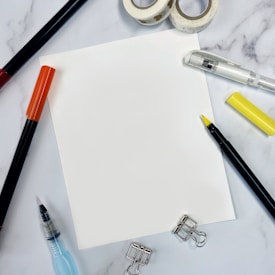 A blank white sheet of paper is centered on a marble surface. Surrounding the paper are various stationery items including a white and a yellow pen, a red marker, a blue water brush pen, two rolls of washi tape with hearts, metal binder clips, and a black marker with a yellow cap.