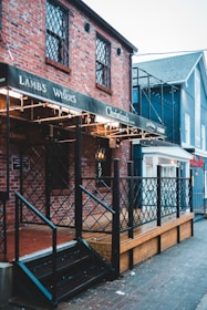 A brick building with decorative lattice windows and a wooden deck features a sign with multiple brand names. The building is adorned with string lights and has a metal railing leading up to a black stairway. Adjacent structures are visible, displaying a mix of brick and painted surfaces.