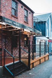 A brick building with decorative lattice windows and a wooden deck features a sign with multiple brand names. The building is adorned with string lights and has a metal railing leading up to a black stairway. Adjacent structures are visible, displaying a mix of brick and painted surfaces.